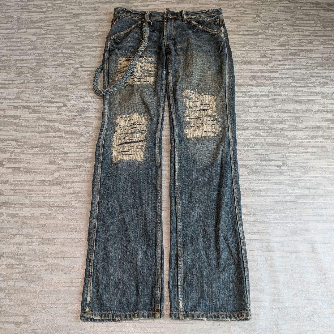 Flat-lay photo of distressed women's denim jeans with ripped knees and a braided chain accessory on a light patterned background.