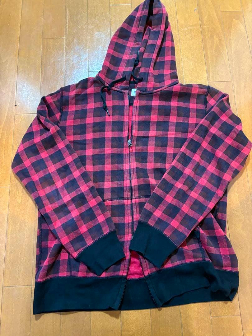 Red and black checkered Uniqlo zip-up hoodie laid flat on a wooden floor, showing the front with hood, zipper, and black cuffs.