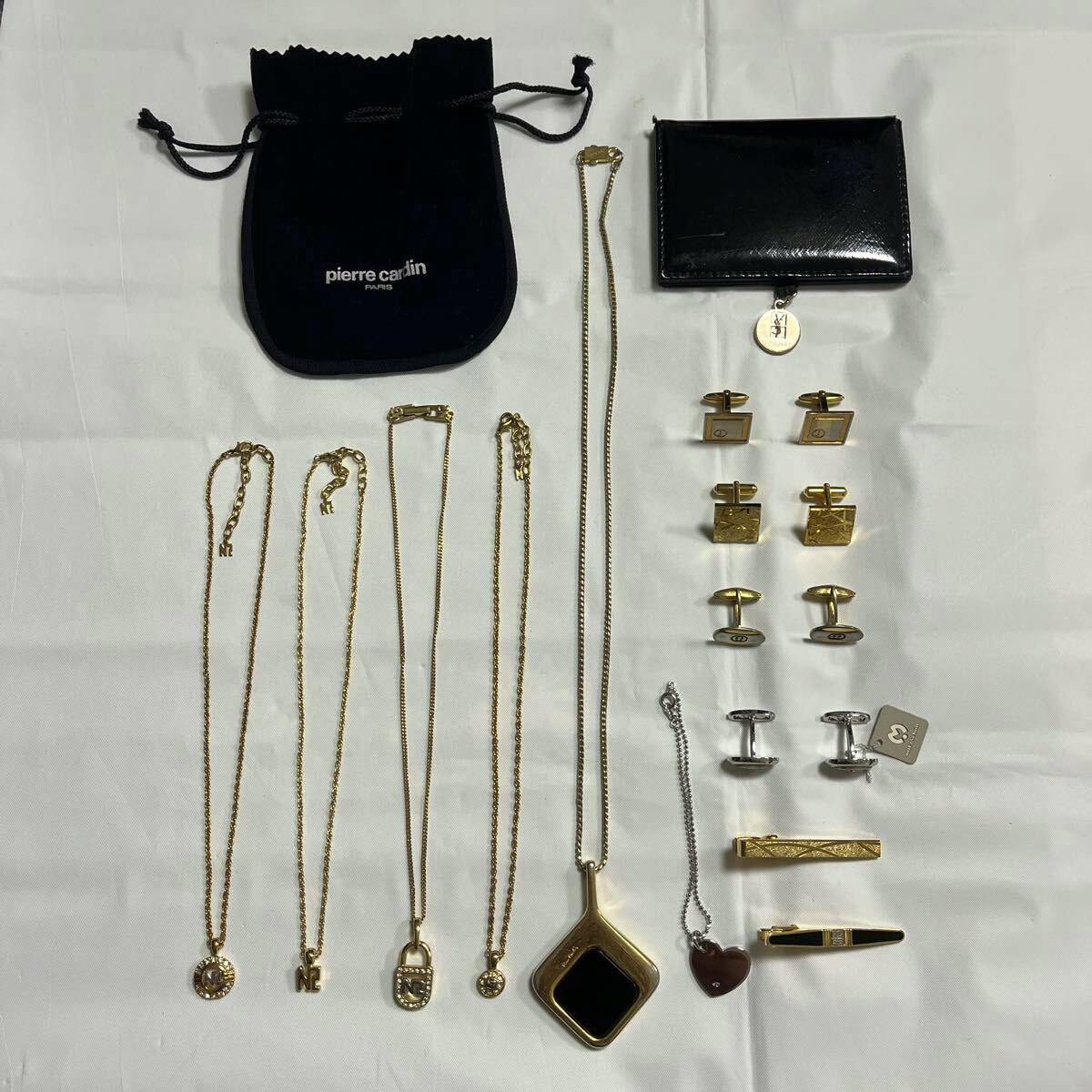Flat lay of multiple necklaces with pendants, bracelets, cufflinks, tie clips, and small pouches arranged on a white background.
