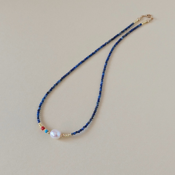 Thin necklace of small lapis lazuli beads with a central freshwater pearl, a small red coral bead, and gold spacer beads shown on a plain background.