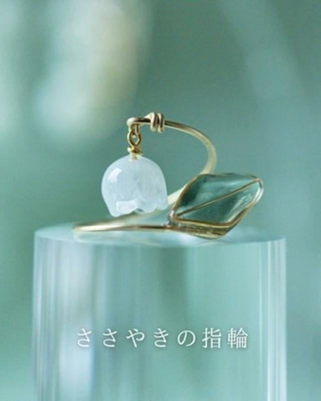 A ring with a small white bell-shaped charm and a green leaf-shaped stone displayed on a clear pedestal.