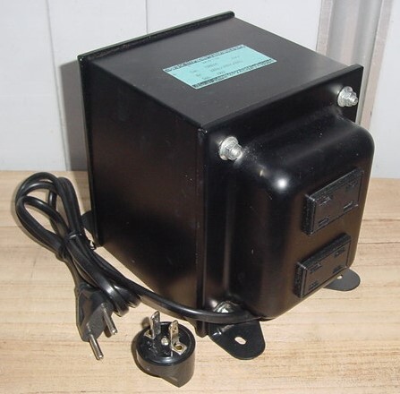 Black metal transformer unit with attached power cord and plug adapters sitting on a wooden surface.