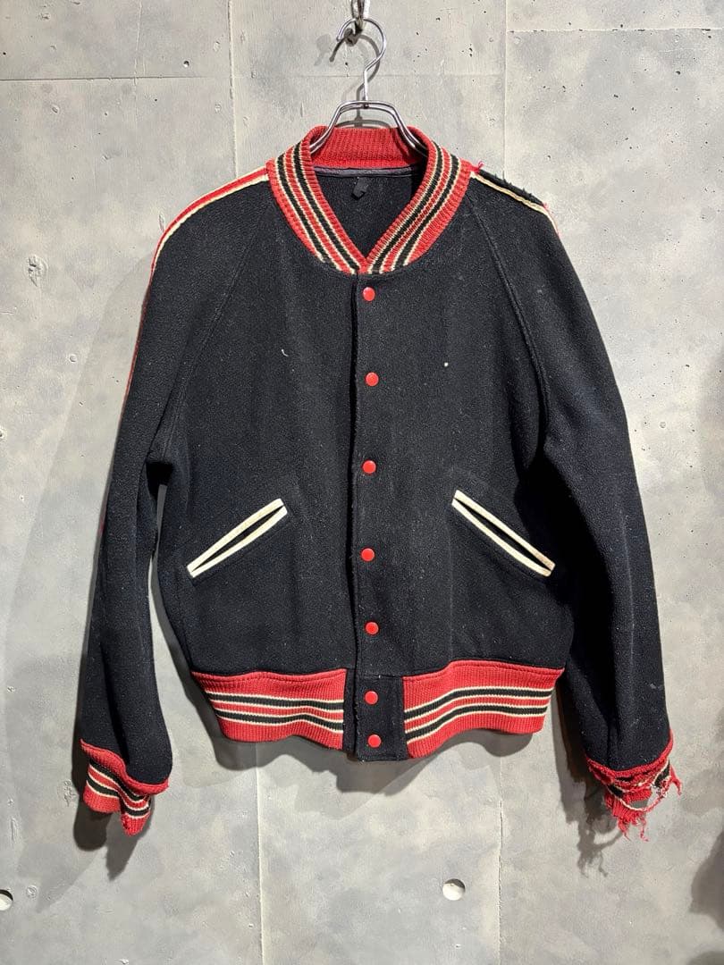 Front view of a black vintage men's varsity-style jacket (スタジャン) with red-striped ribbing, red snap buttons, and white-trimmed pockets hanging on a hook.