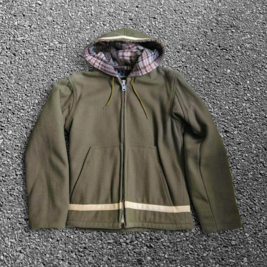 Flat-lay of a men's wool parka with a hood showing plaid lining, full zipper, and front pockets on a textured background.