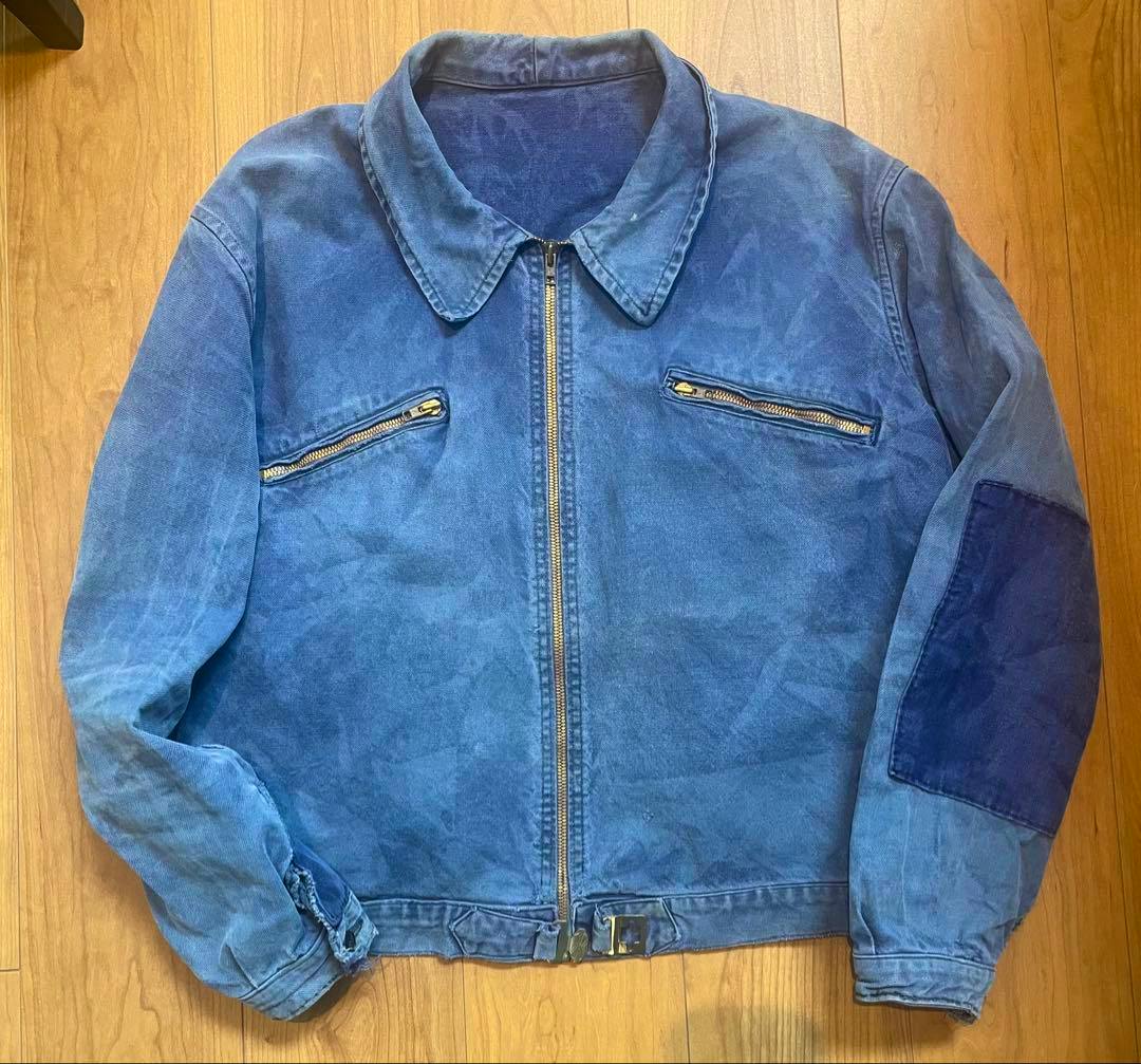 Front view of a blue vintage men's cotton cycling jacket laid flat showing a central zipper, two zip chest pockets, and an elbow patch on the sleeve.