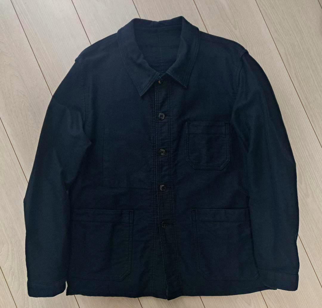 Front view of a black moleskin French work men's jacket laid flat on a wooden floor, showing buttons and three pockets.