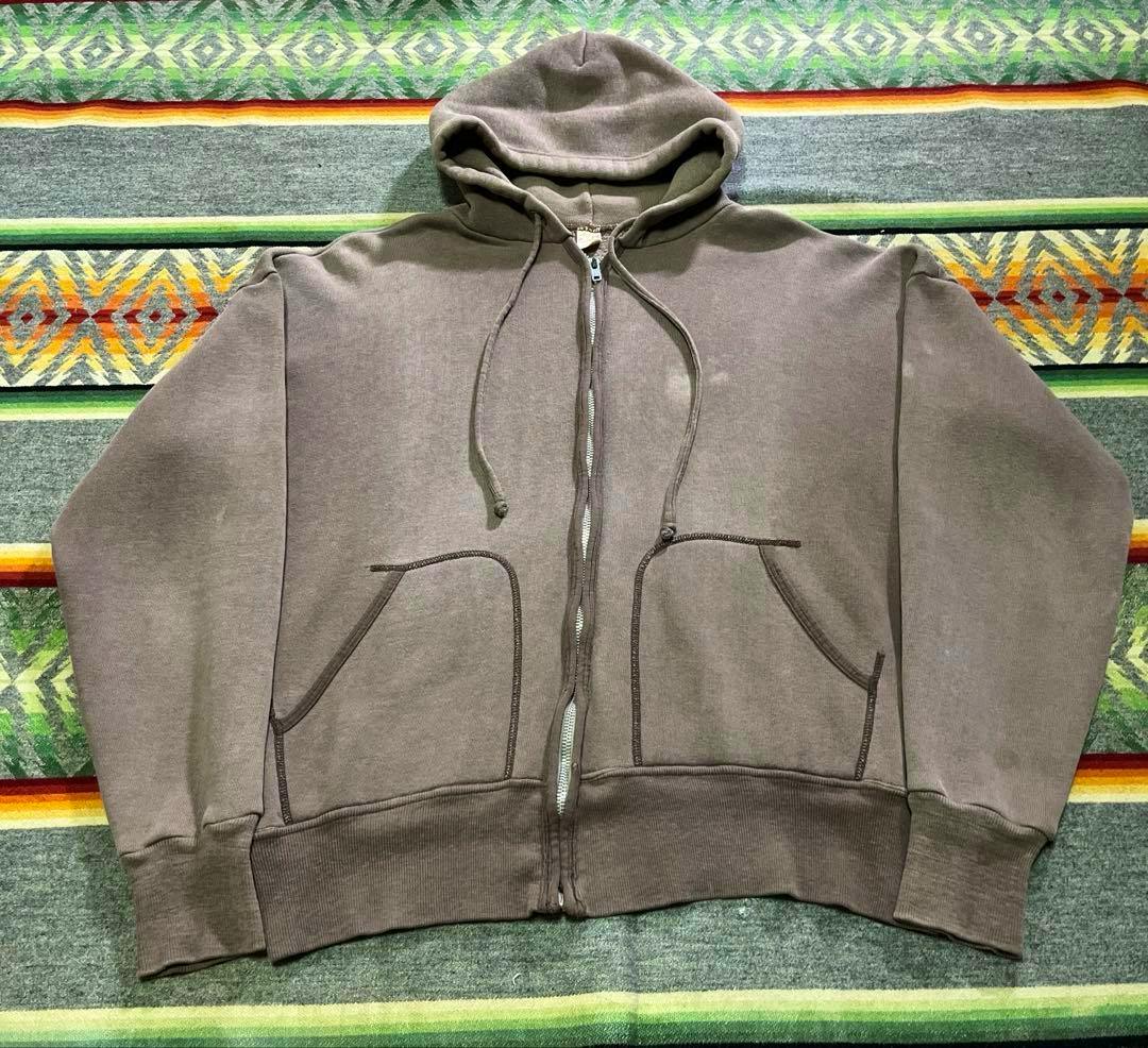 Brown zip-up hooded cotton sweater laid flat on a patterned blanket showing the front, zipper, drawstrings, and kangaroo pockets.