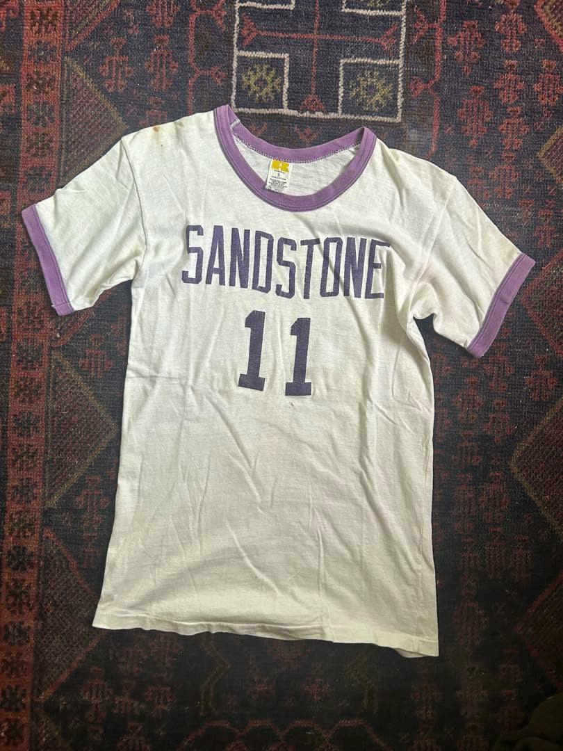 Front view of a white ringer T-shirt with purple collar and sleeve bands and "SANDSTONE 11" printed on the chest.