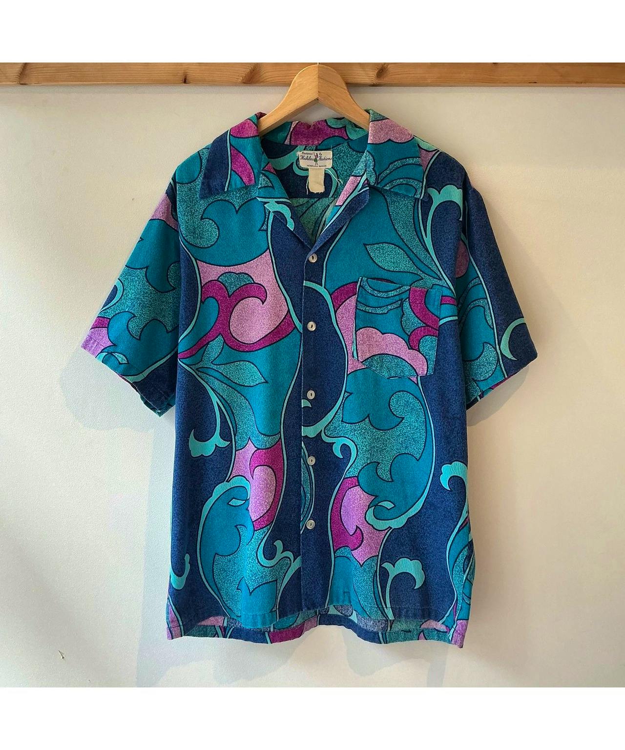 Front view of a blue and purple vintage Hukilau Fashions men's cotton Hawaiian short-sleeve open-collar shirt hanging on a wooden hanger.