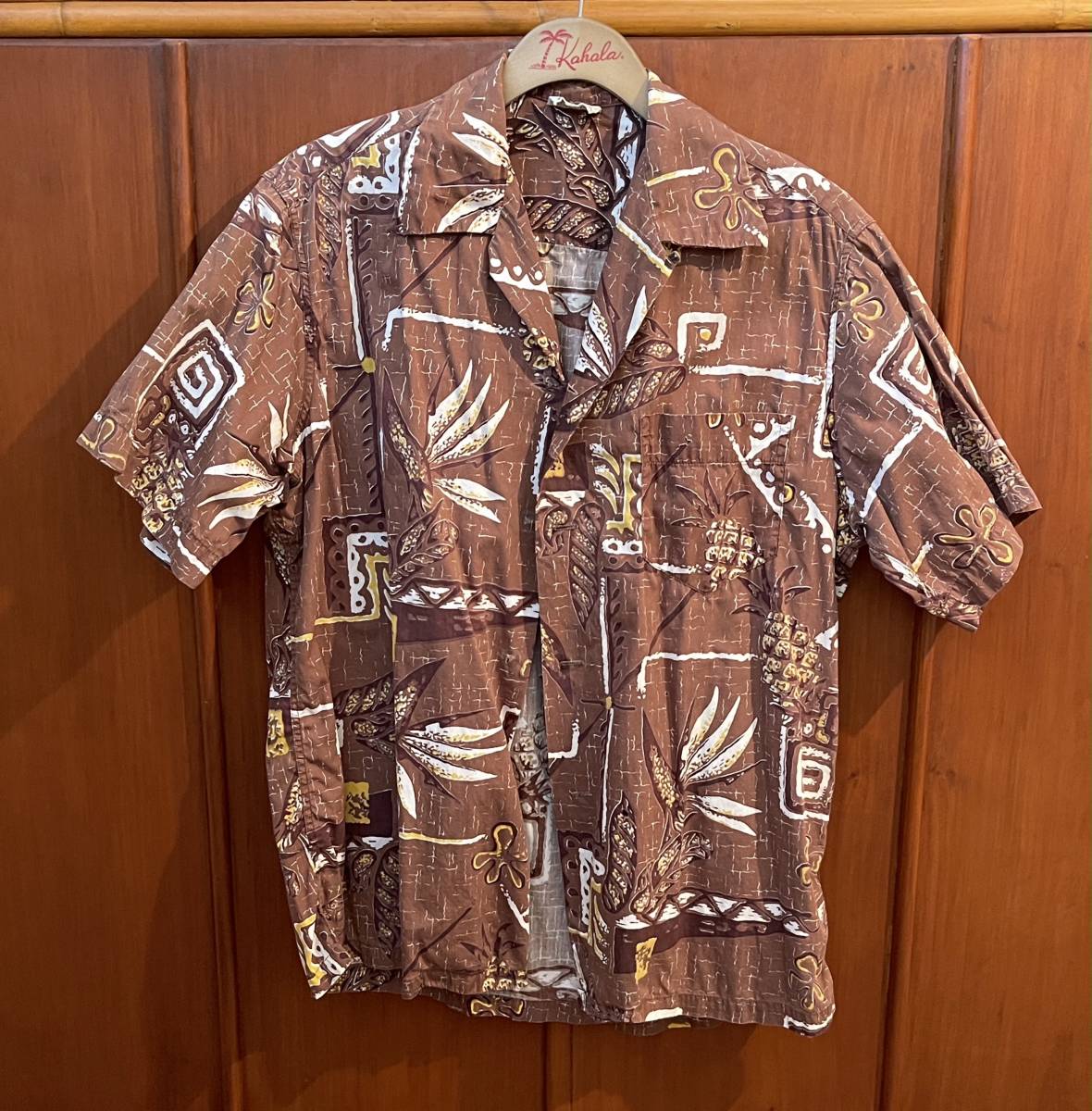 Front view of a brown 1960s Shaheen's of Honolulu vintage men's cotton aloha shirt (size L) with abstract botanical and geometric print hanging on a wooden hanger against a wood background.