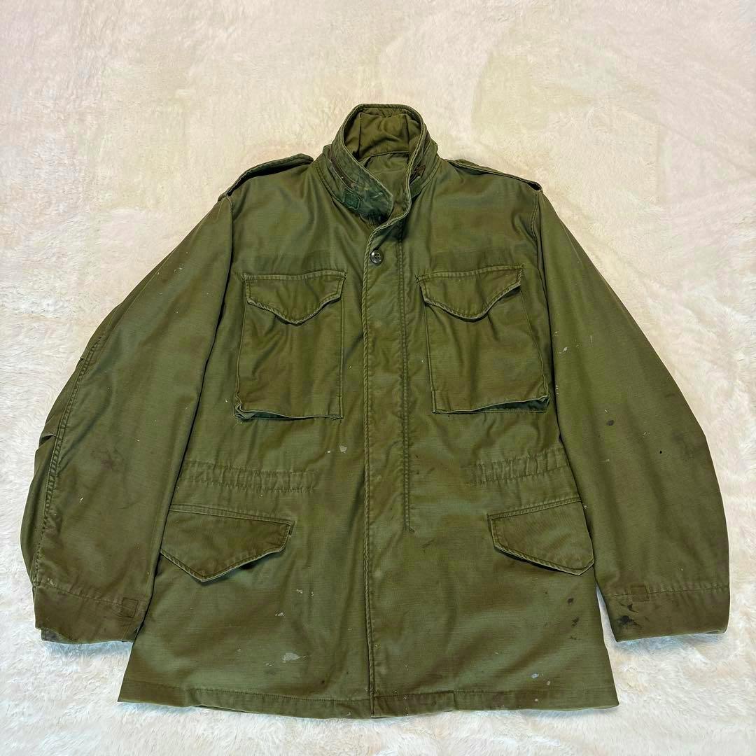 Olive-green M65 field jacket laid flat showing front pockets and paint stains.