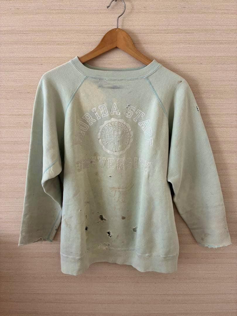 Front of a light green vintage cotton sweatshirt with a faded "Florida State University" print and multiple holes, hanging on a wooden hanger.