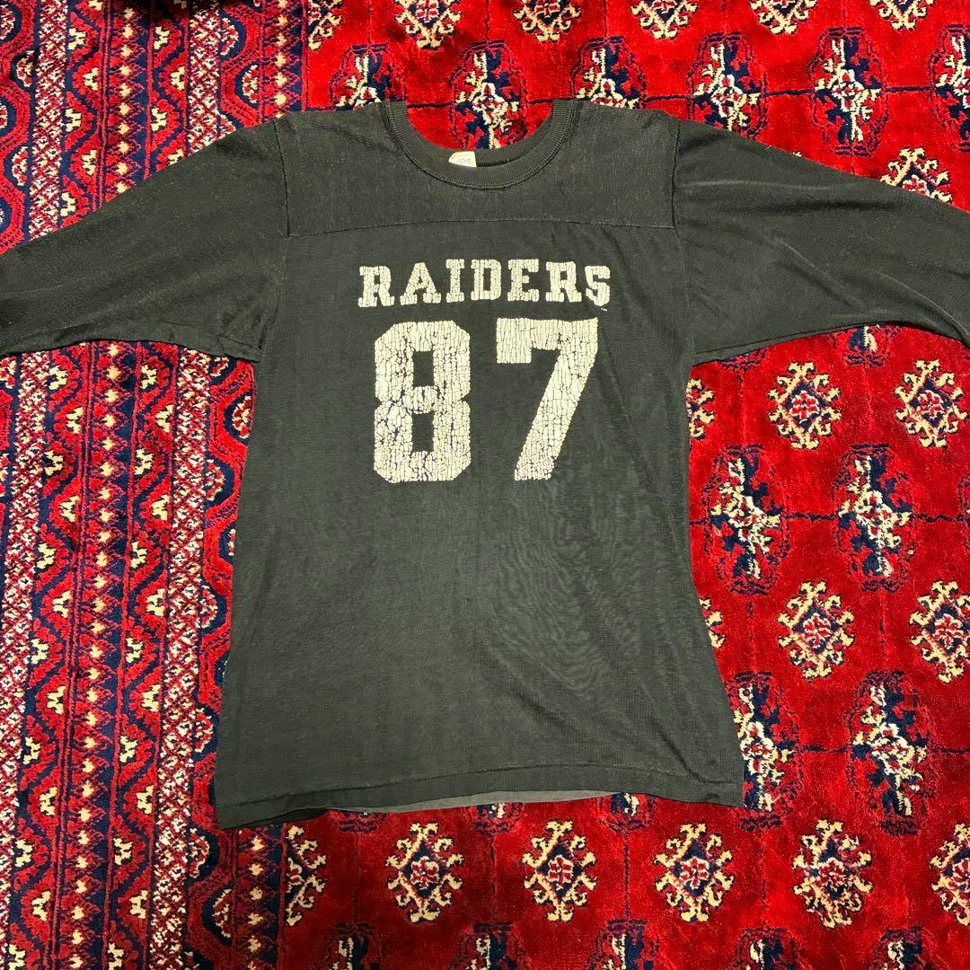 Black vintage Raiders T-shirt with cracked white "RAIDERS 87" print laid flat on a red patterned rug.