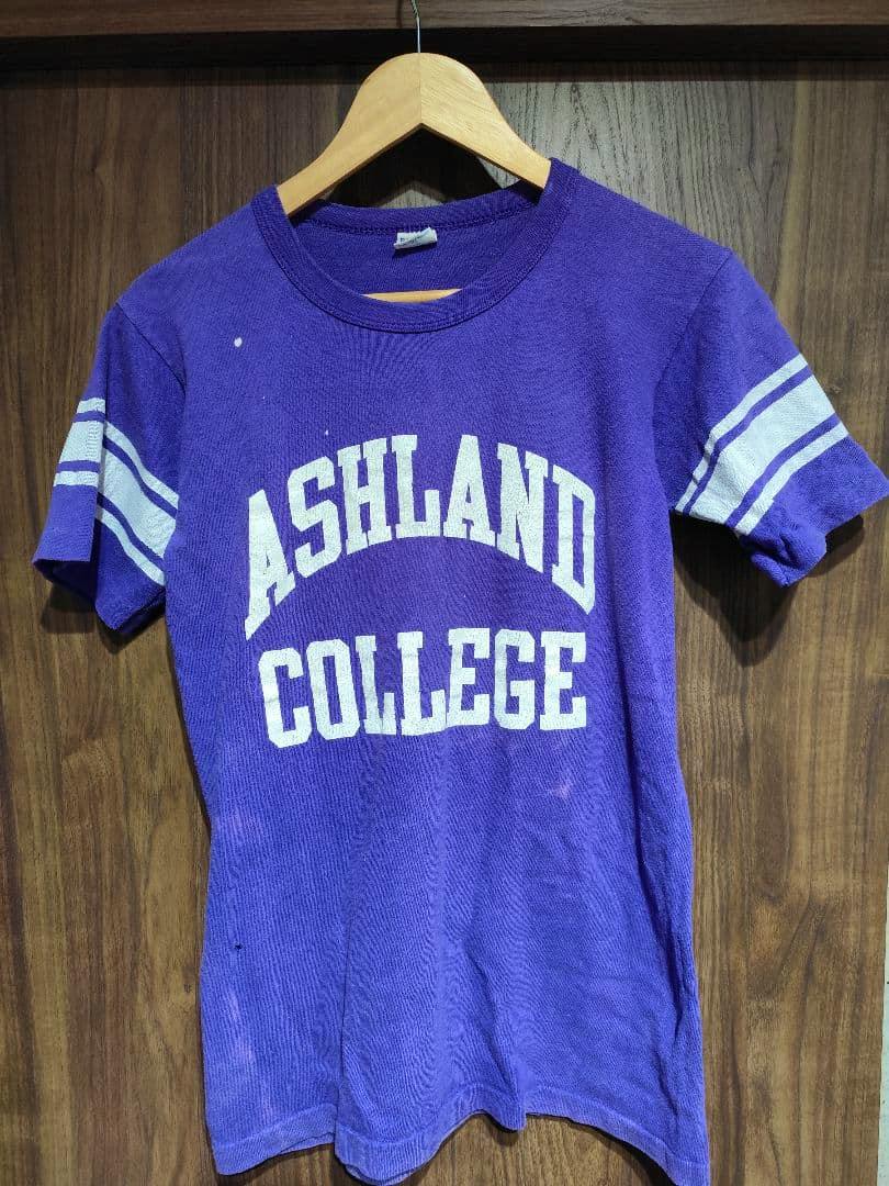 Purple men's football T-shirt with "ASHLAND COLLEGE" printed on the front and white sleeve stripes, hanging on a wooden hanger.