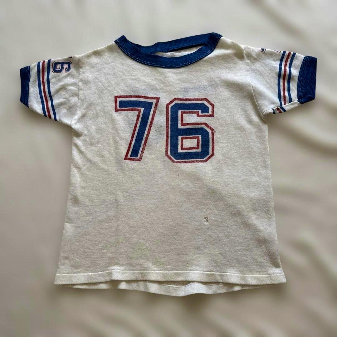 White vintage ringer men's T-shirt laid flat showing blue-and-red "76" numbering on the front with blue ribbed collar and sleeve cuffs.