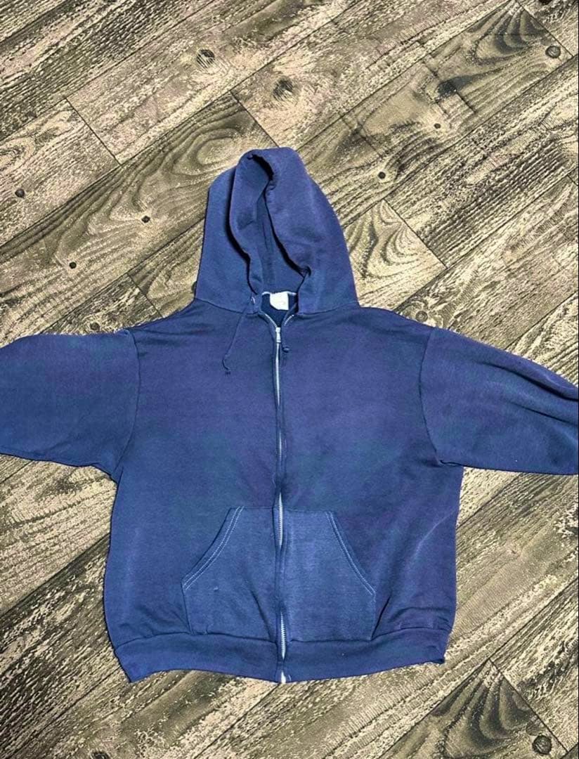Front view of a blue zip-up hoodie laid flat on a wood-pattern floor, showing the hood, zipper, and front kangaroo pocket.
