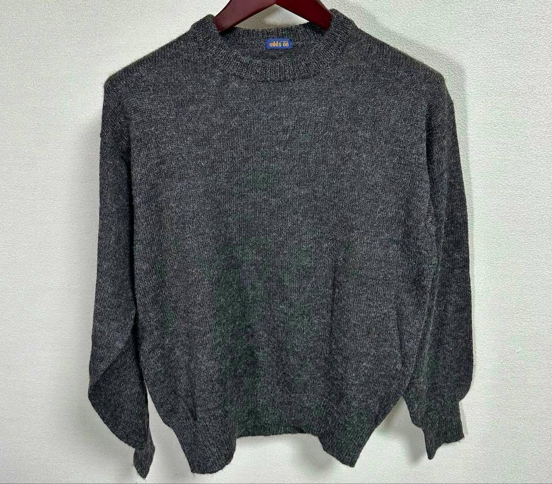 Front view of a dark gray wool sweater (Issey Miyake "odds on") hanging on a hanger against a white wall.