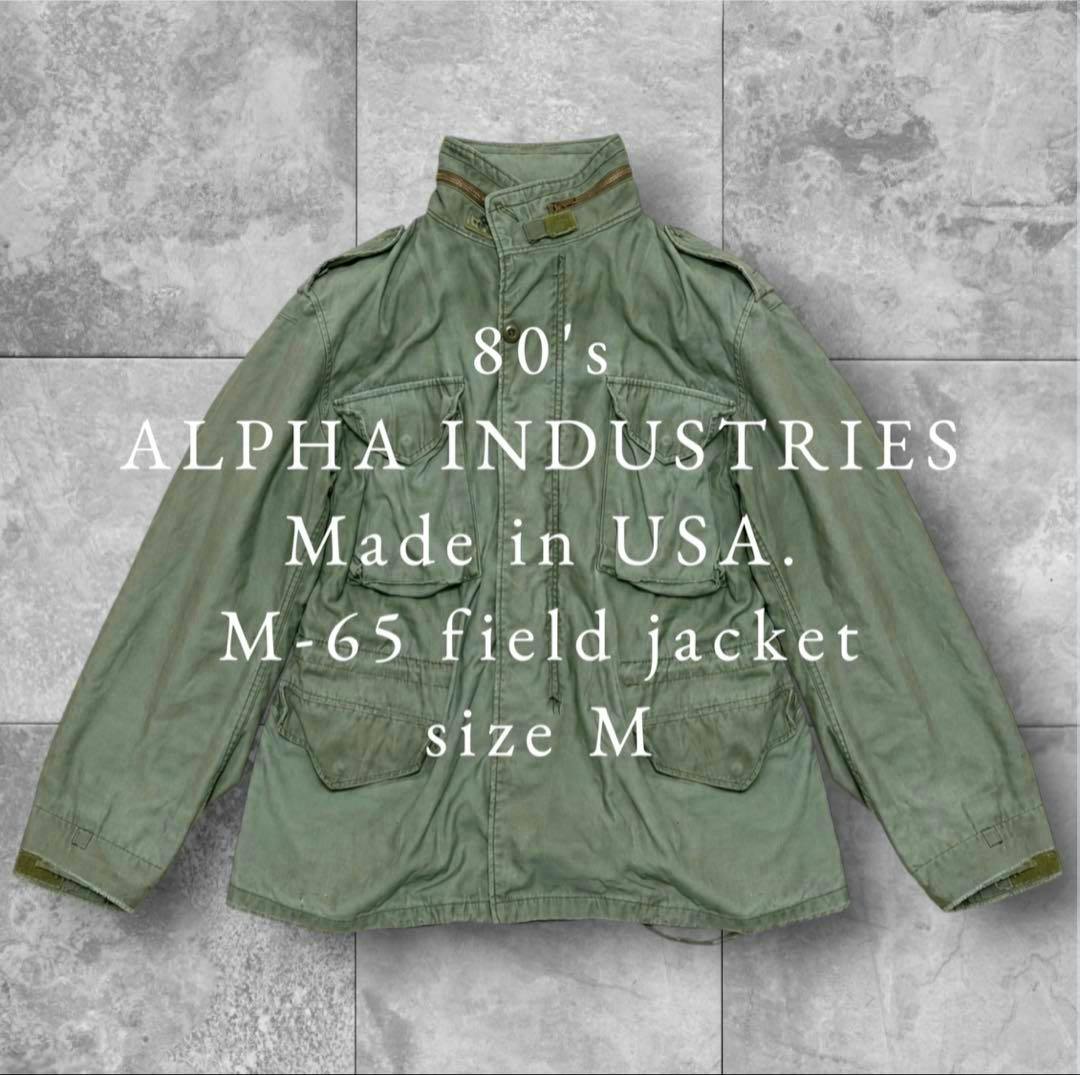 Front view of a green M-65 field jacket laid flat with white text overlaid on the image.