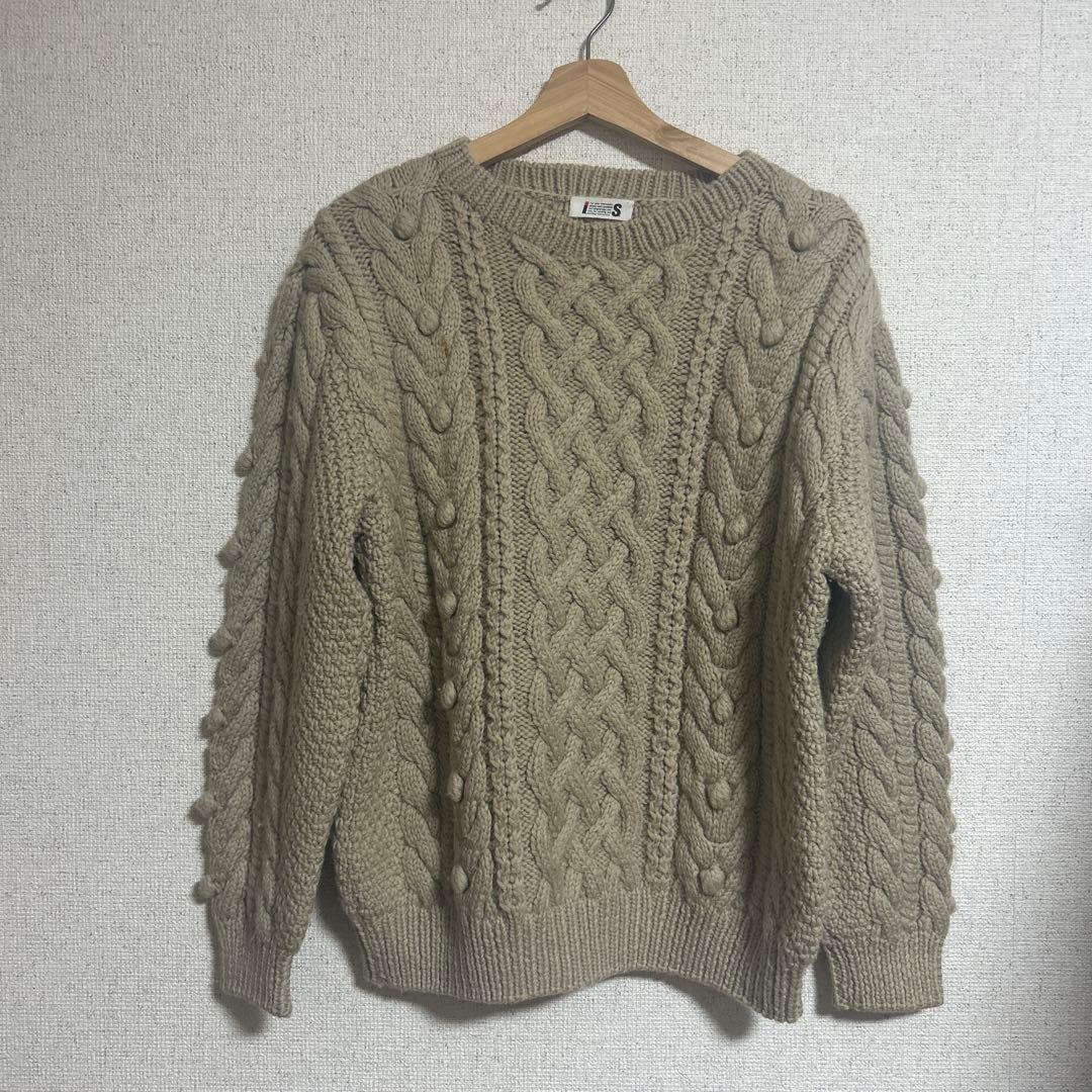 Front view of a hand-knitted wool sweater (1980s I.S. issey sport) hanging on a wooden hanger against a textured wall.