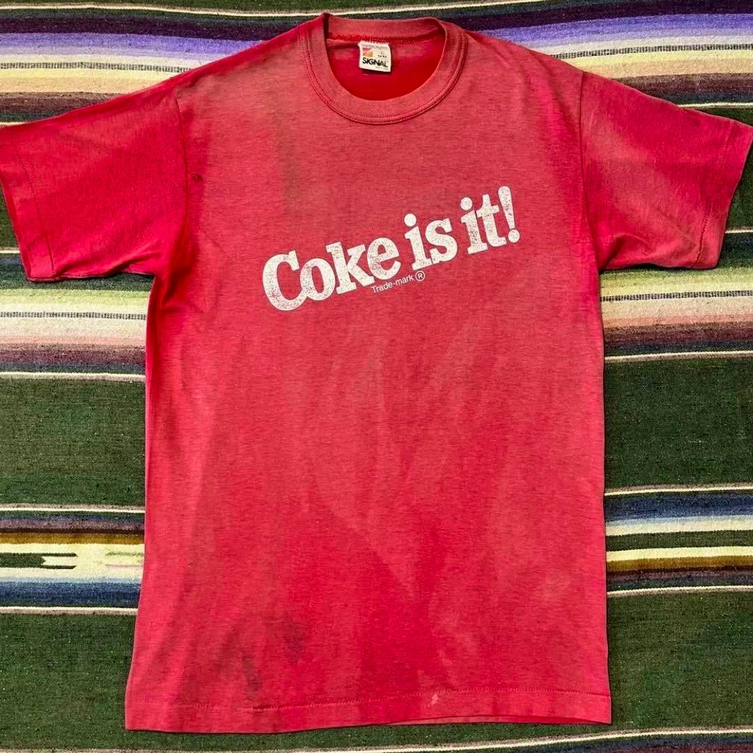 Red Coca‑Cola T‑shirt laid flat showing a white "Coke is it!" chest print, overall fading, and a small hole near the left sleeve.