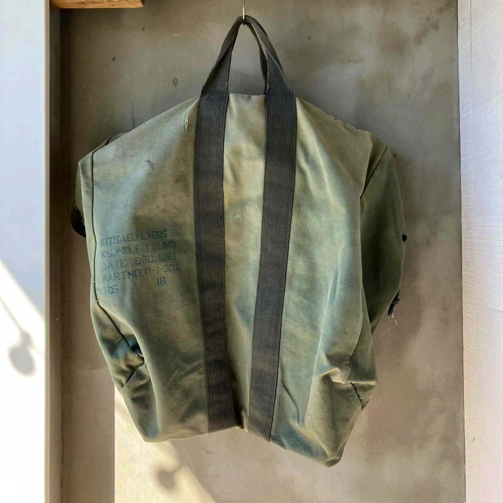 Green canvas USAF flyers kit bag hanging on a hook, showing two black carrying straps and stenciled text on the side.