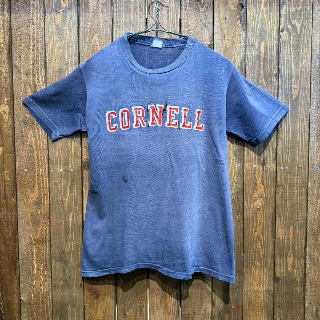 Front view of a faded blue Cornell printed T-shirt hanging on a hanger against a wooden background.