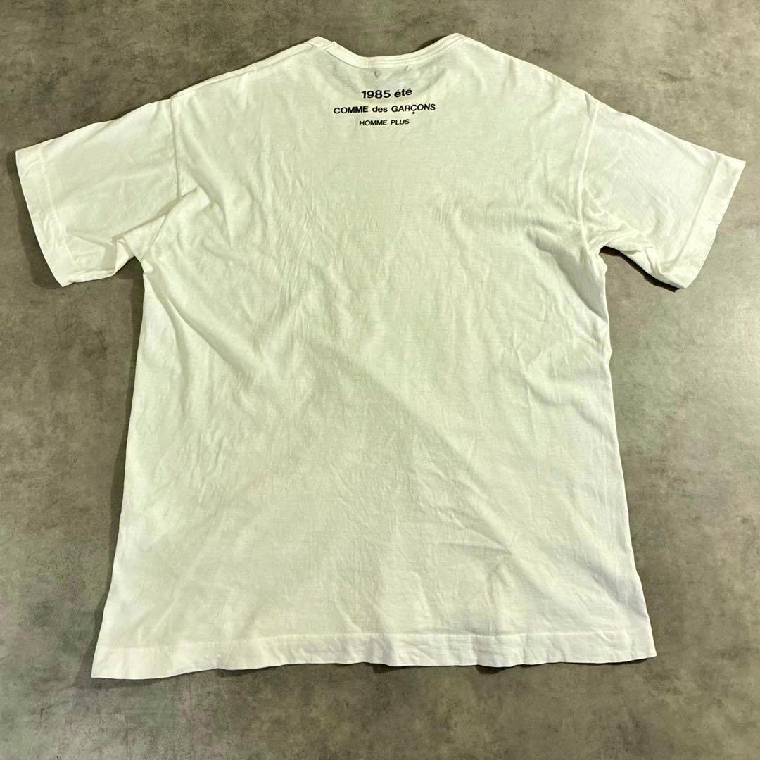 White cotton T-shirt laid flat showing the back with small "1985 été" and "COMME des GARÇONS HOMME PLUS" printed near the neckline.
