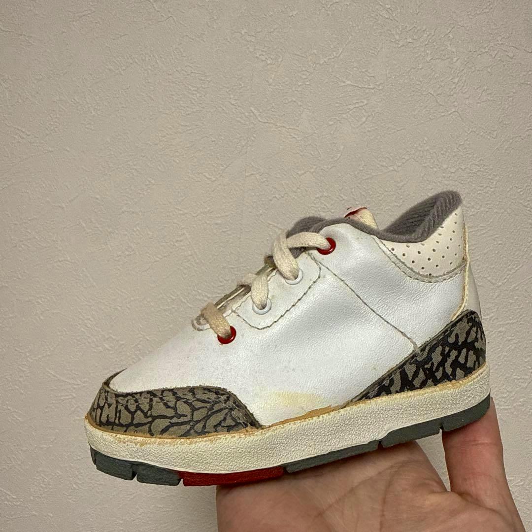 A baby Jordan 3 sneaker held in a hand showing side profile with white leather and elephant-print accents.