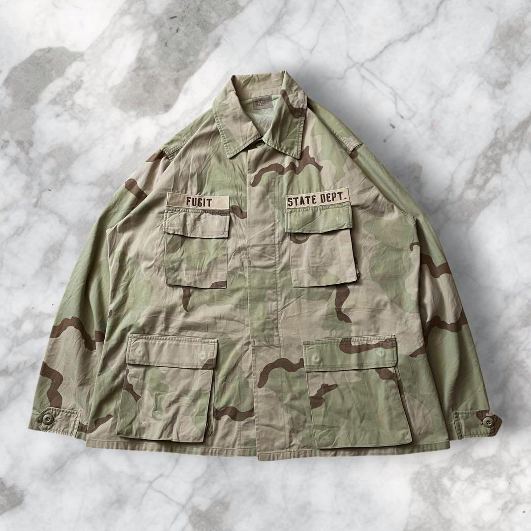 Front view of a vintage US military BDU jacket in desert camouflage laid flat, showing chest patches labeled “FUGIT” and “STATE DEPT.” and four front pockets.