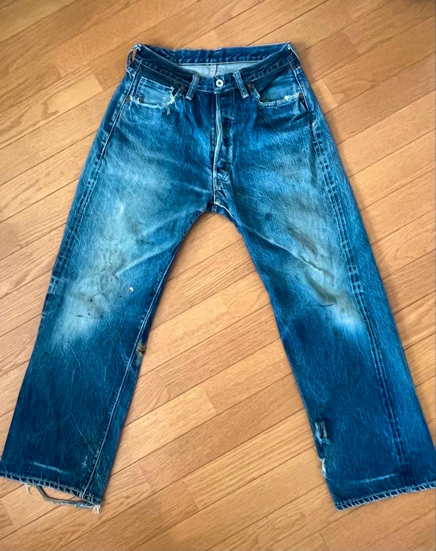 Flat-lay front view of men's blue denim jeans with faded areas, paint stains, and frayed hems on a wooden floor.