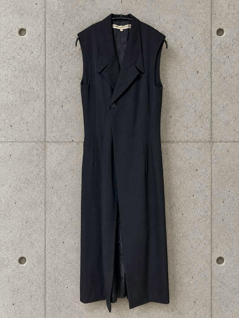 Front view of a long black sleeveless wool vest/dress hanging on a hanger against a concrete wall.