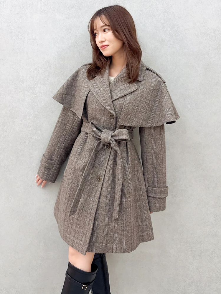 Model wearing the 2-way cape mini trench coat in brown checked fabric, belted at the waist, standing against a gray wall.