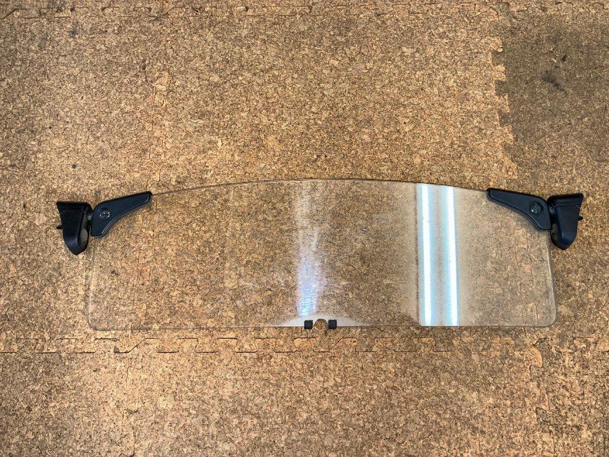 Transparent wind deflector with black mounting brackets shown laid flat on a cork surface.