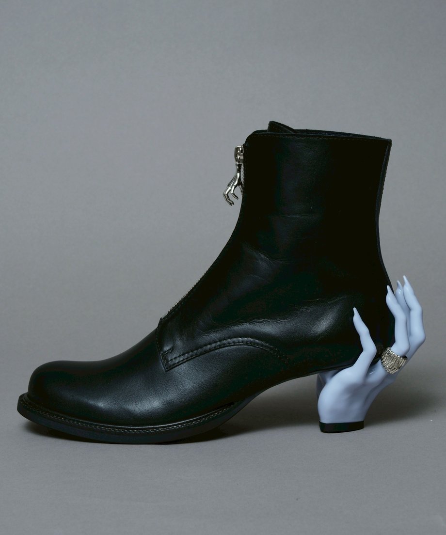 A black leather ankle boot photographed in profile with a pale sculpted hand forming the heel.
