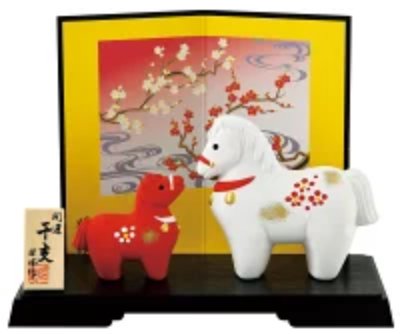 Two ceramic horse figures (one white and one red) displayed on a black stand with a gold folding screen backdrop and a small wooden plaque.