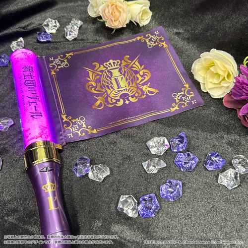 Photograph of the Veil of Order official multicolor pen light lit purple, shown with its decorative sleeve/box and scattered clear and purple gem props on a black surface.