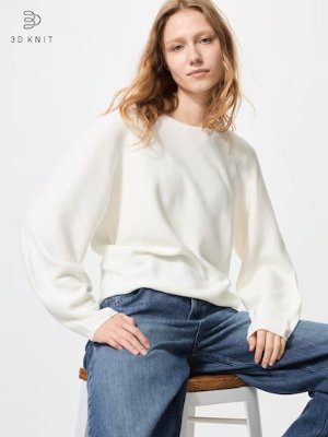 Model wearing a white cotton crew-neck sweater with volume sleeves, seated on a stool.