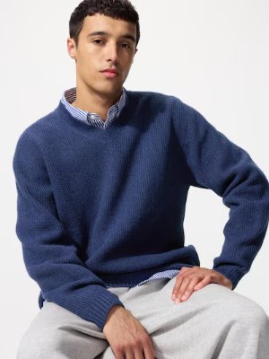 A seated male model wearing a blue knit sweater layered over a collared shirt.
