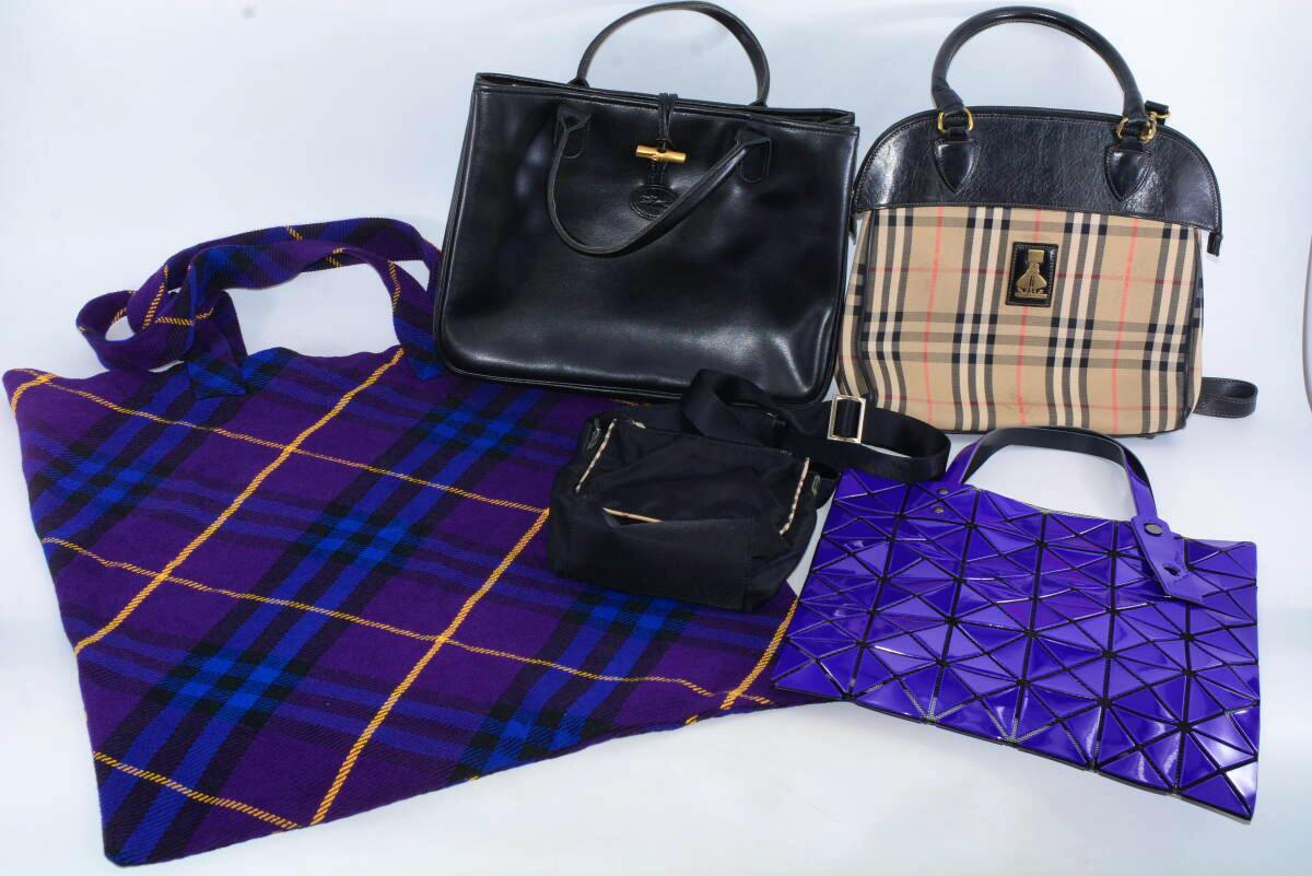A group of five bags on a white background: a purple plaid fabric tote, a black leather handbag, a Burberry check handbag, a small black pouch, and a purple geometric Issey Miyake-style tote.