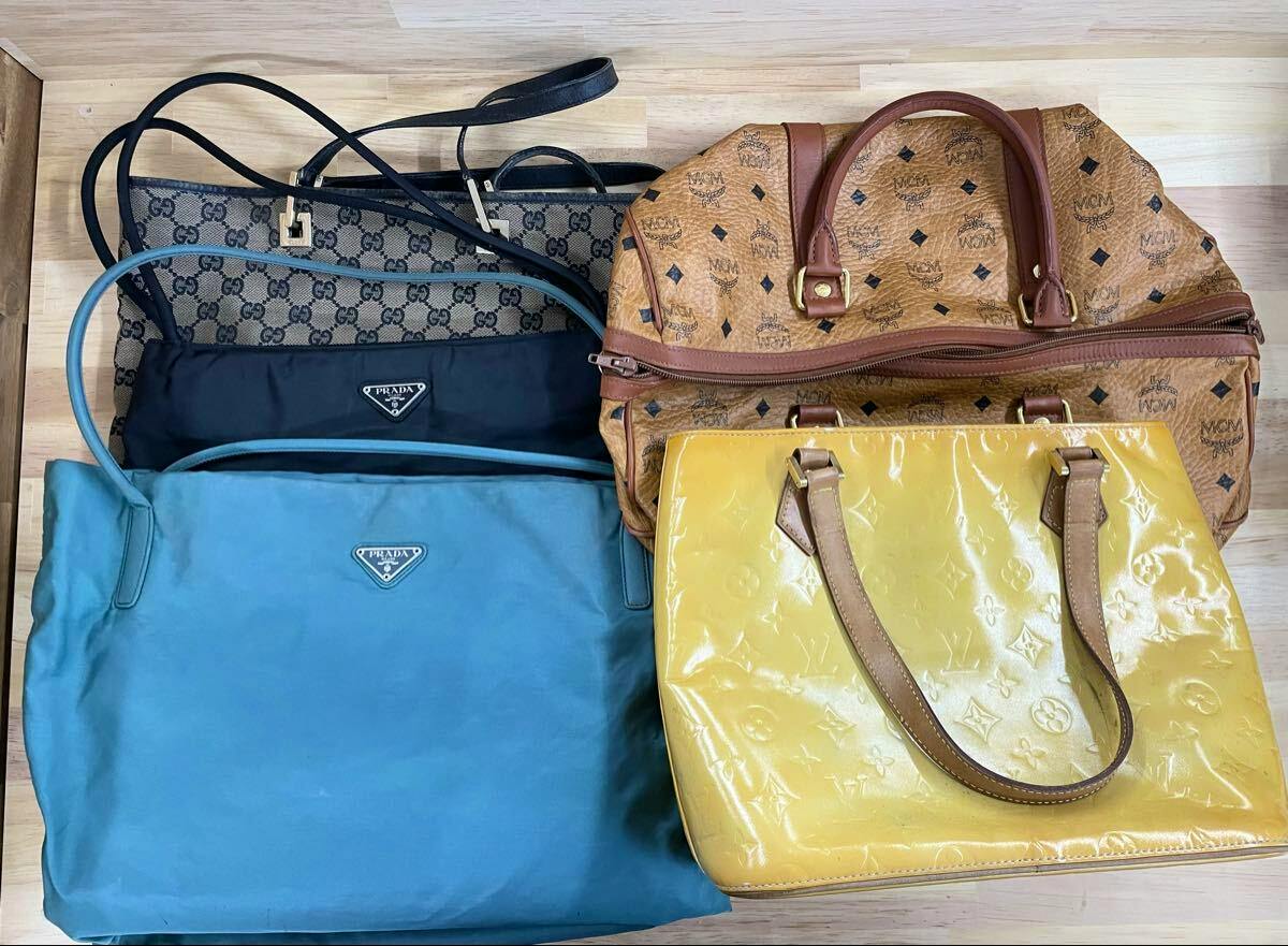 Top-down photo of five brand bags including a yellow Louis Vuitton bag and blue and black Prada bags arranged with other designer bags on a wooden surface.