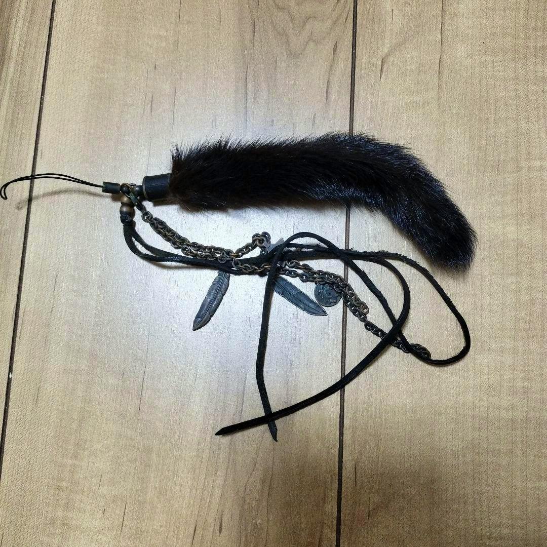 A dark fur charm strap with metal chains, feather pendants, and leather cords laid on a wooden floor.