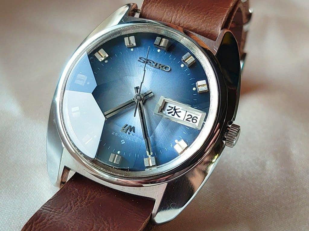 Close-up photograph of the watch showing a blue faceted dial, day/date window, and brown leather strap.