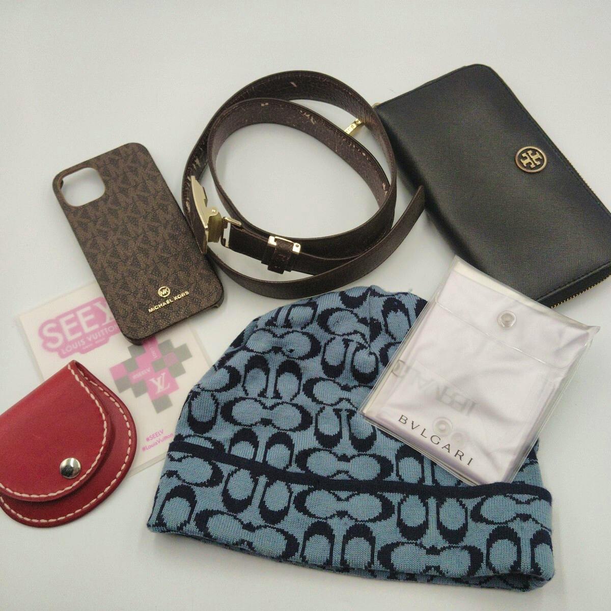 Flat lay of assorted designer accessories and leather goods including a belt, black zip wallet, patterned knit hat, phone case, and small pouches.
