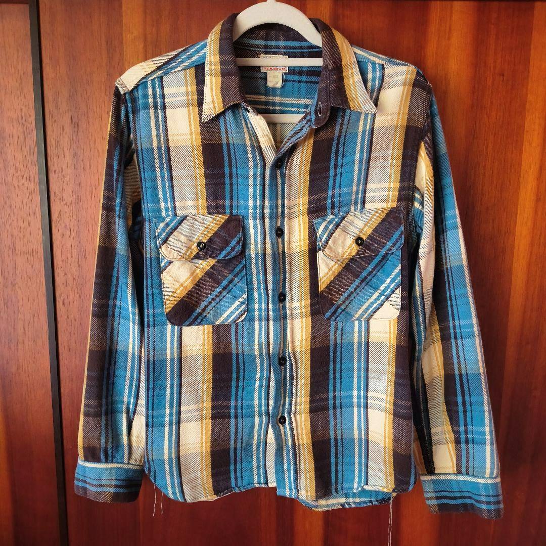 Front view of a men's cotton plaid flannel shirt (blue, yellow, cream, and brown) hanging on a hanger against a wooden background.