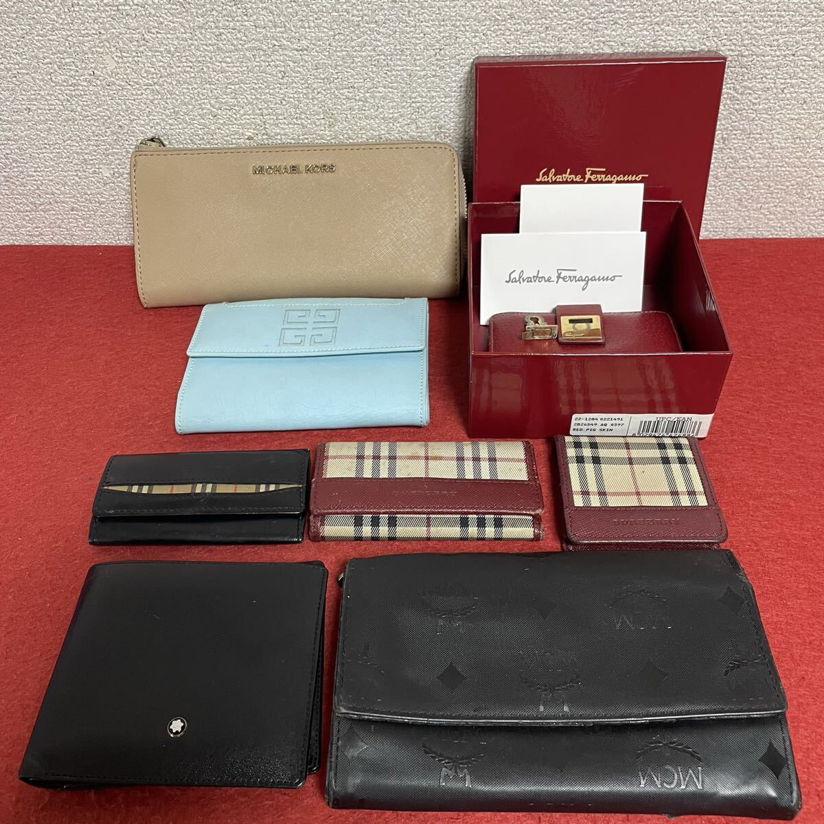 Eight branded wallets and small leather goods arranged on a red surface, including a beige zip-around wallet, Burberry check pieces, an MCM embossed wallet, a Montblanc item, and a Ferragamo box.