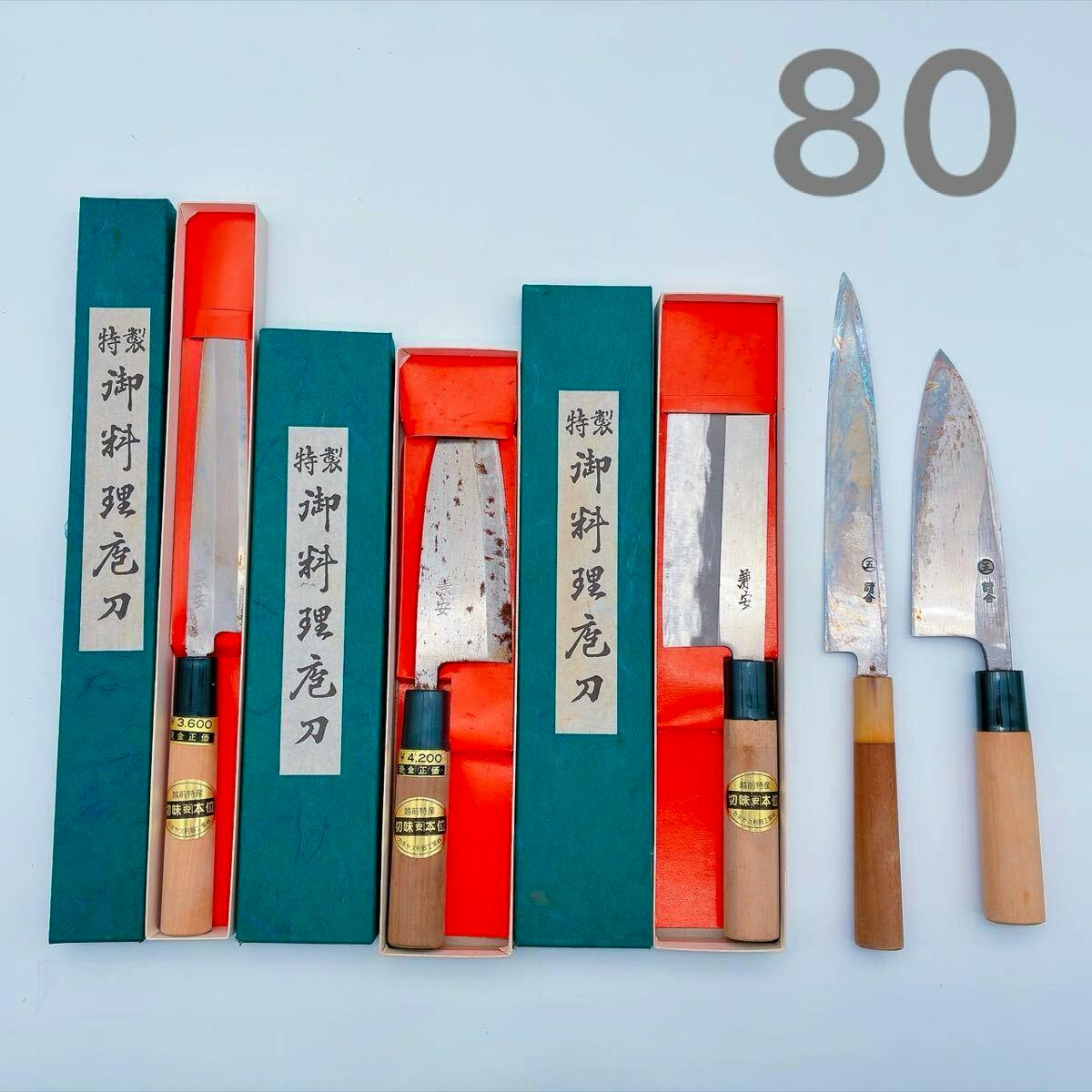 A set of Japanese chef knives displayed with their green boxes with red interiors, and three loose knives arranged on a pale background.