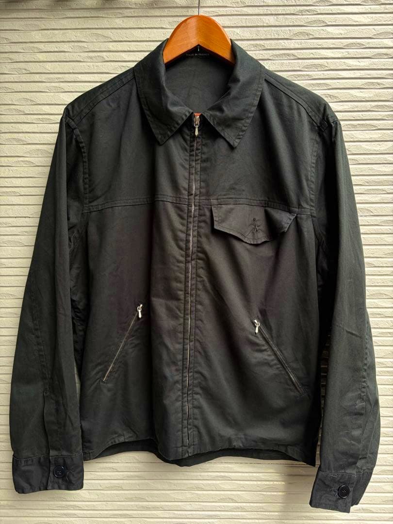 Front view of the black men's cotton Agnes b zip jacket hanging on a wooden hanger against a textured wall.