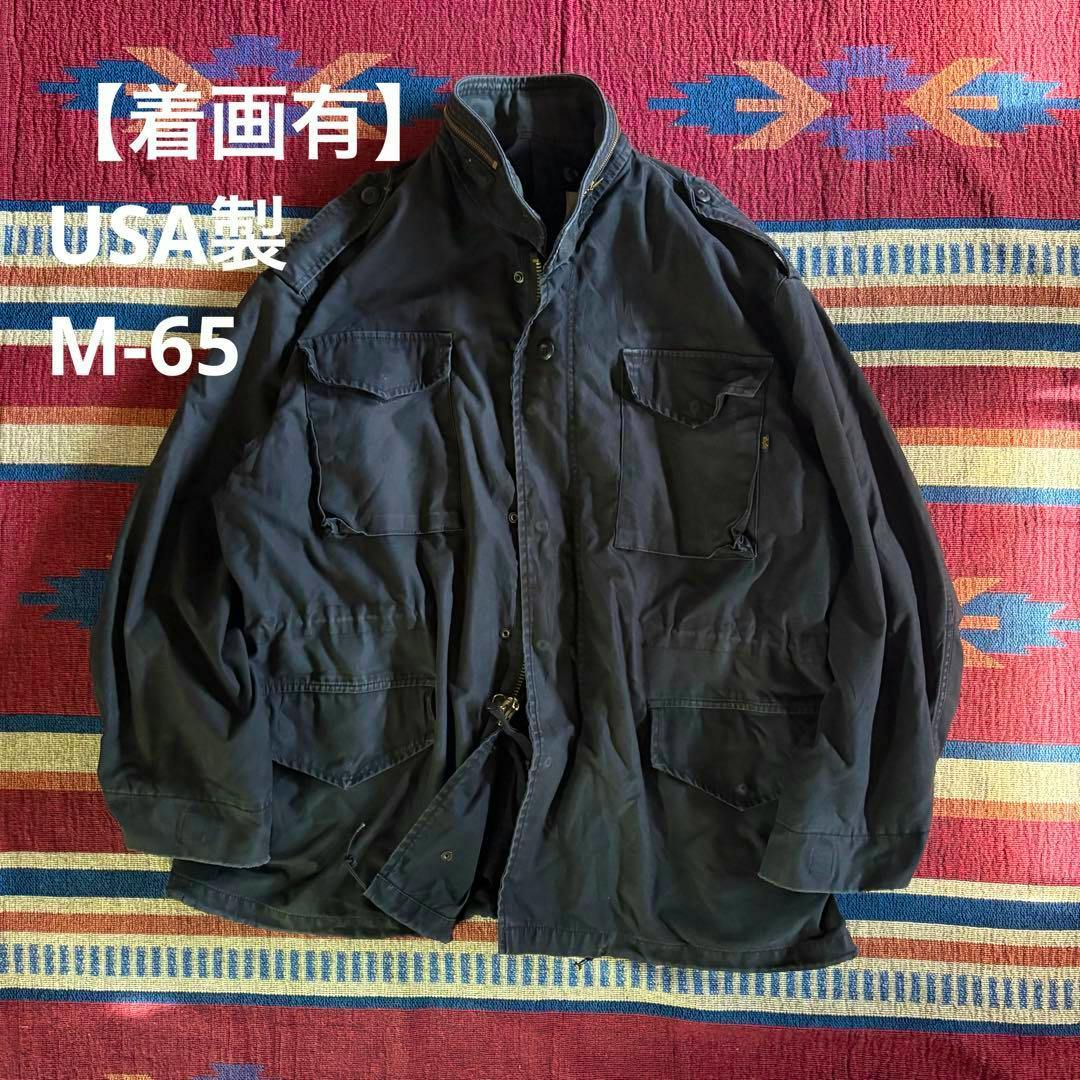 Black USA-made M-65 field jacket laid flat on a patterned rug, showing front pockets and the zipper.