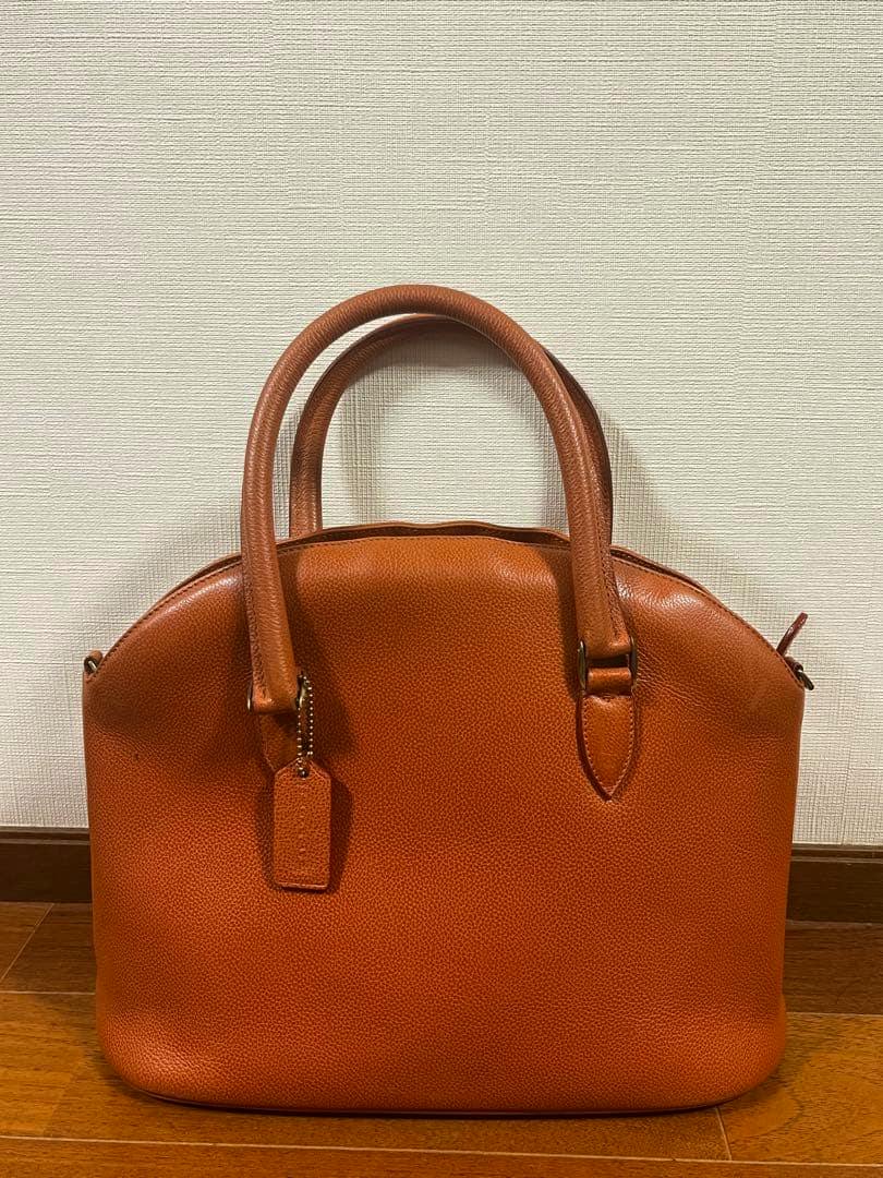 Front view of a brown cow leather 90s Coach handbag with double handles and a leather hang tag against a plain wall.