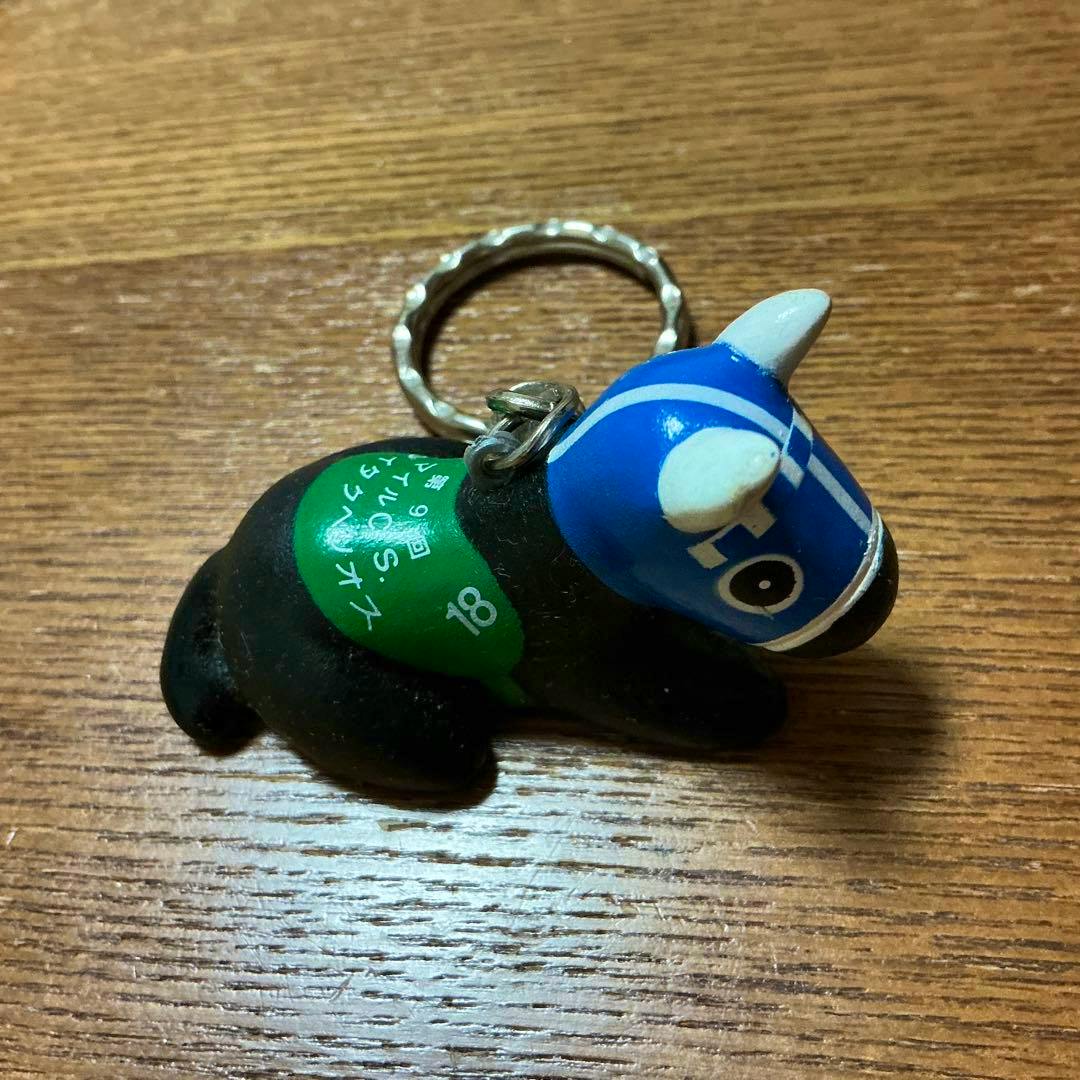 A horse-shaped key holder/figure with a blue mask, green saddle, and metal key ring on a wooden surface.